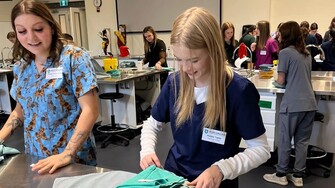 a student wearing scrubs fold cloths in a medical setting. More students and staff in scrubs are in the background.