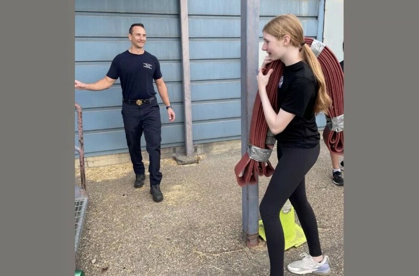 A student carries a folded fire hose over her shoulder while a firemedic looks on.